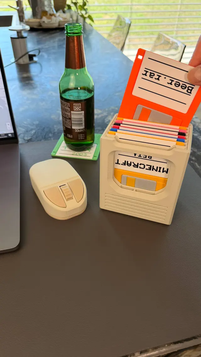 Mouse beige stampato in 3D su una scrivania accanto a una bottiglia di birra e a un box di floppy